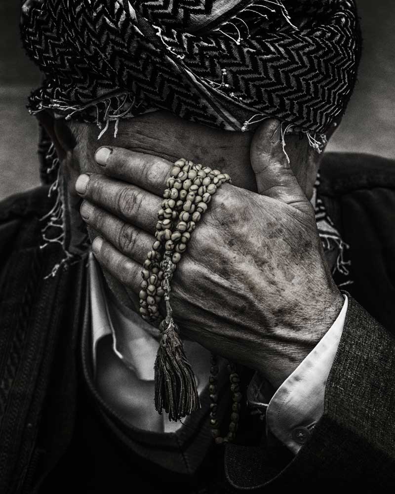 An old man wearing a turban with prayer beads wrapped around his hand places his hand over his face in a gesture of shame.